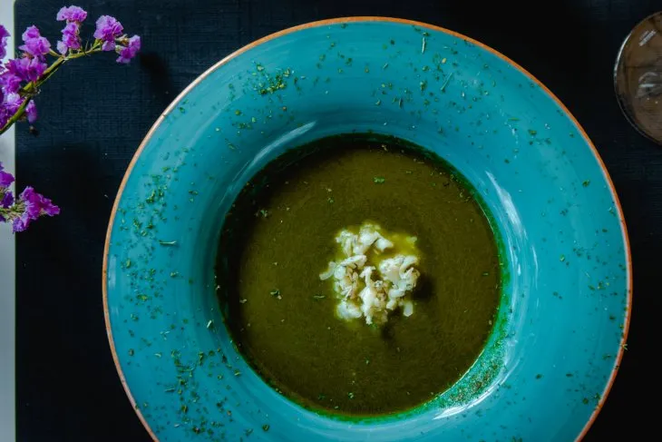  a plate of green pumpkin soup with gorgonzola cheese in the middle, purple flowers on the left 